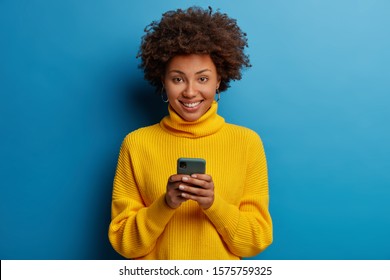 Photo of dark skinned lady enjoys distant communication, uses mobile phone, surfes fast unlimited internet, has pleasant smile, dressed in yellow sweater, makes shopping online, isolated on blue