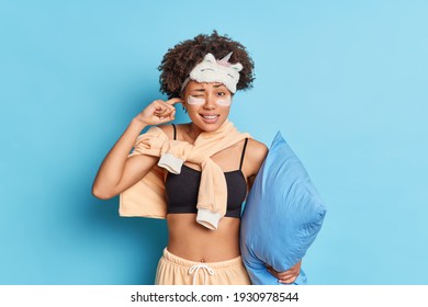 Photo Of Dark Skinned Woman Plugs Ears Cannot Fall Asleep Because Of Noise Wears Nightwear Sleeping Mask Applies Patches Under Eyes Holds Pillow Isolated Over Blue Background. Sleep Interruption