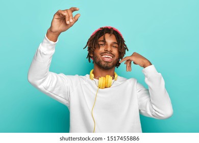 Photo Of Delighted Happy Teenager With Dreadlocks, Raises Arms, Feels Joy As Listens Favourite Music Via Headphones, Moves In Rhythms Of Song, Wears White Jumper, Poses Against Blue Background