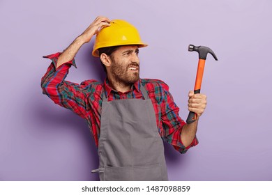 Photo of discontent handyman turns right, looks into distance with frowned face, holds hammer, being professional builder, notices new object for repairing wears helmet, apron. Renovation, engineering