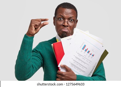 Photo Of Displeased Dark Skinned Business Owner Studies Information Data In Documents, Demonstrates Small Gesture, Dissatisfied With Low Company Income, Isolated Over White Background. Too Tiny