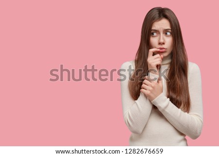 Photo of displeased sad girlfriend looks with frustrated expression aside, keeps hand near lips, has worried look dressed in white jumper models against pink studio wall with empty space for your text