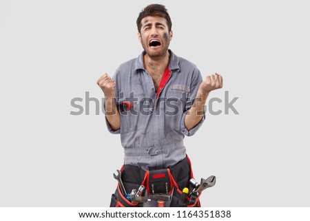 Photo of downcast stressed engineer clenches fists desperately, being despondent by bad work conditions, feels tired of constant hard labour, dressed in casual clothes, poses against white background