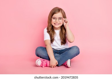 Photo of dream funny little girl sit floor wear glasses white t-shirt jeans shoes isolated pink color background