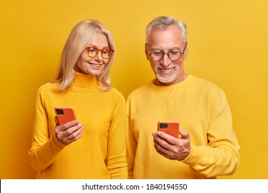 Photo of elderly grandmother and grandfather view photos together on smartphone devices watch interesting funny video online dressed in casual yellow turtlenecks pose indoor. Age and technology