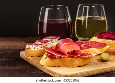 A photo of embutidos tapas, sandwiches with jamon, salchichon, lomo, and other Spanish sausages, with glasses of wine and green olives, on a dark background with a place for text