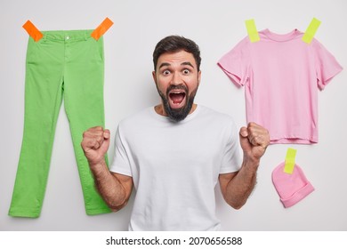 Photo of emotional bearded young man clenches fists roots for someone watches game eclaims loudly poses against white background with plastered clothes behind. People emotions reactions concept