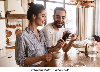 Photo of european brunette couple man and woman 20s drinking coffee and using cell phone during breakfast in kitchen at home - Powered by Shutterstock