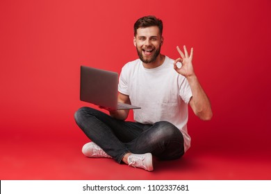 Photo Of European Joyful Guy In T-shirt And Jeans Sitting On Floor With Legs Crossed And Showing Ok Sign While Holding Laptop Isolated Over Red Background