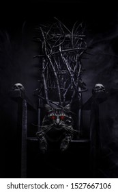 Photo Of Evil Cat With Glowing Eyes On Witch Throne Made Of Branches And Skulls On Foggy Black Background.