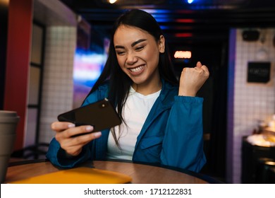 Photo Of Excited Asian Woman Playing Video Game On Mobile Phone And Making Winner Gesture While Sitting In Cafe