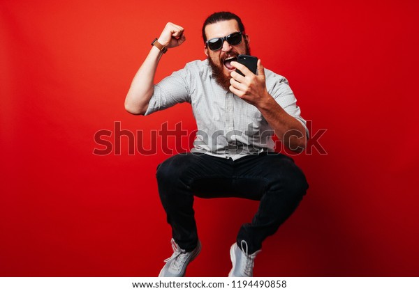 赤い背景に携帯電話を使用して飛び跳ねる 興奮した髭の男の写真 の写真素材 今すぐ編集