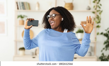 Photo Of Excited Black Woman Singing To Her Smart Phone At Home, Listening To Music In Wireless Earphones