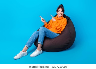 Photo of excited cheerful lady wear knitted pullover sitting bean bag showing two fingers empty space isolated blue color background
