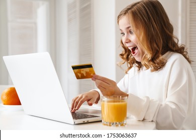 Photo Of Excited Cute Girl Using Laptop And Holding Credit Card While Drinking Juice In White Kitchen