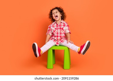 Photo Of Excited Funny School Girl Dressed Checkered Clothes Sitting Green Chair Looking Empty Space Laughing Isolated Orange Color Background