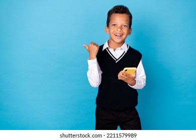 Photo Of Excited Positive Little Guy Dressed Black Vest Pointing Thumb Empty Space Chatting Modern Device Isolated Blue Color Background