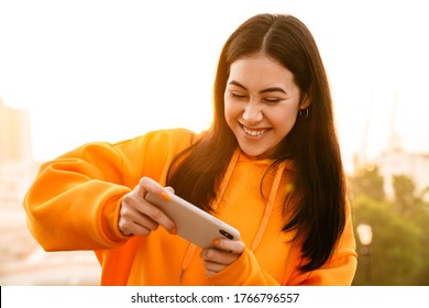 Photo Of Excited Pretty Asian Woman Playing Video Game On Cellphone While Walking Outdoors