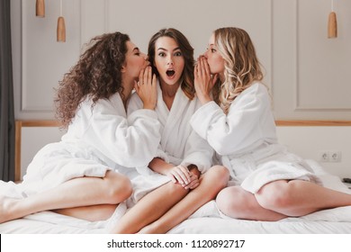 Photo Of Excited Three Women 20s Wearing Bathrobe Sitting On Big Bed And Telling Gossips To Each Others During Bridal Shower In Posh Apartment Or Fashionable Hotel Room