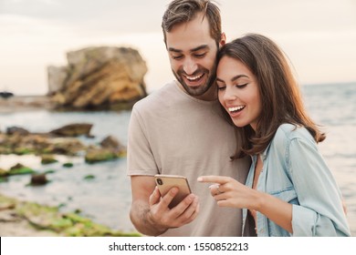 Photo of excited young couple wearing casual clothes using and pointing finger at cellphone while walking on sunny beach - Powered by Shutterstock