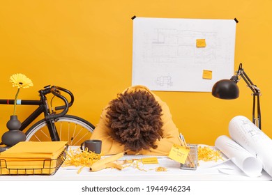 Photo of exhausted tired woman leans on table worked all day on architectural project wants to sleep poses at desktop with rolled paper sketches stickers. People deadline occupation concept.