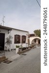 Photo of exterior of a house in rural Greece, with white walls and brown shutters, a small garden with plants and flowers, the entrance to a restaurant with an outdoor seating area, on a sunny day 