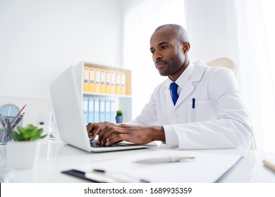 Photo Of Family Doc Dark Skin Guy Watch Modern Technology Notebook Novelty Innovation Medical Reform Cov Viral News Wear White Lab Coat Sitting Chair Table Office Clinic Indoors