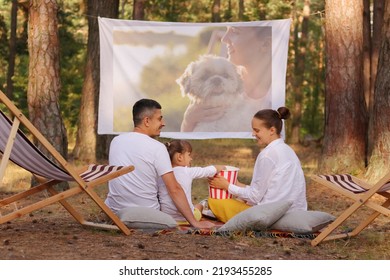 Photo Of Family Sitting In The Forest On Deck Chairs With Their Little Daughter And Watching Movie On Projector, Parents Looking At Each Other With Smile, Family Enjoying Time Spending Together.