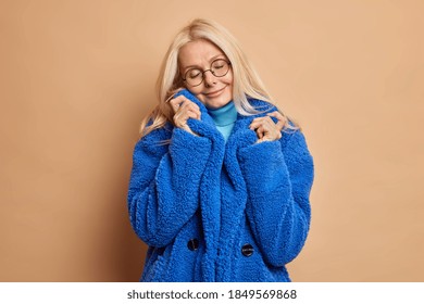 Photo of fashionable blonde woman tilts head and closes eyes wears round spectacles blue fur coat recalls something pleasant isolated over brown background. Winter fashion and style concept.