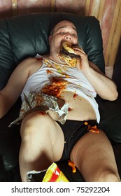 Photo of a fat couch potato eating a huge hamburger and watching television.  Harsh lighting from the television illuminates the dark room.