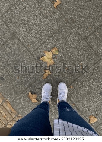 Photo of female man standing on paving stones in white sneakers and navy blue ripped jeans. Photo taken from above while walking. street photography lifestyle. dry leaves