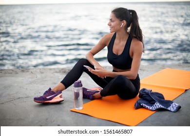 Photo of fit young woman sitting on orange yoga mat outdoors by the sea, holding the phone in one hand, listening to music on wireless headphones, looking away and smiling, wearing black sports wear