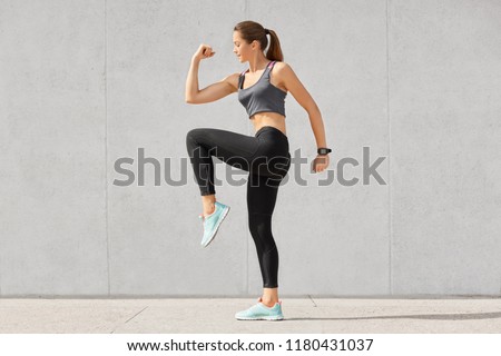 Photo of fitness woman has intense workout, raises legs, dressed in sportsclothes, preapres for running or jogging, poses against grey background. People, exercises, training, lifestyle concept