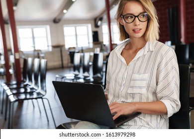 Photo of focused blonde woman wearing eyeglasses working on laptop while sitting in conference hall