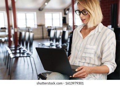 Photo of focused blonde woman wearing eyeglasses working on laptop while sitting in conference hall