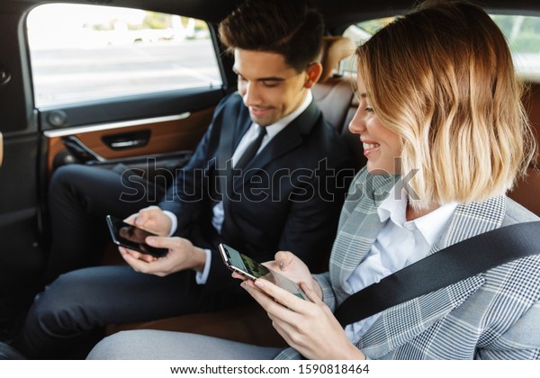 Photo of focused smiling\
colleagues man and woman in formal wear using cellphones while\
going in car