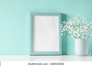 Photo frame and small white flowers in vase on pastel blue background. White gypsophila flowers on shelf or desk. Mock up with decor elements.

