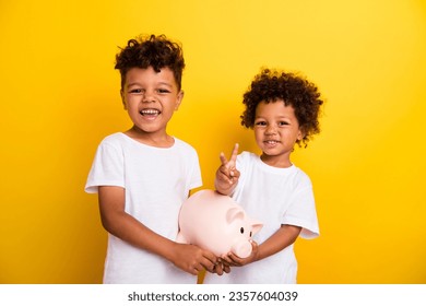 Photo of friendly cheerful little kids hold money bank pig demonstrate v-sign isolated on yellow color background