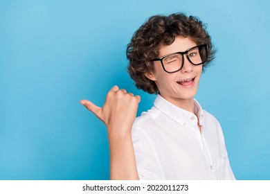 Photo Of Friendly Funny School Boy Wear White Shirt Glasses Winking Pointing Thumb Empty Space Smiling Isolated Blue Color Background