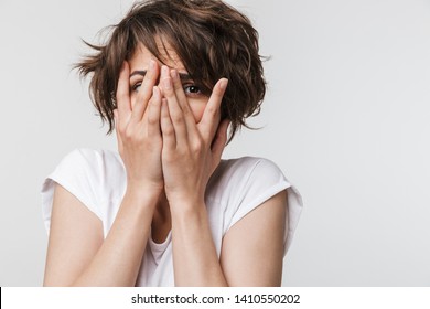 Photo of frightened woman with short brown hair in basic t-shirt keeping hands over her face and looking at camera isolated over white background