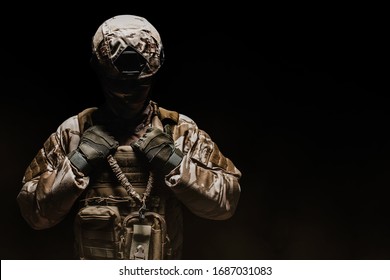 Photo of a fully equipped desert camouflage soldier in mask, helmet, armor and gloves standing on black foggy background front view.