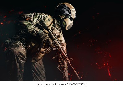 Photo of a fully equipped soldier in armored vest, helmet, face glasses and protection kneeling and attacking with rifle on dark background with ashes flying.
