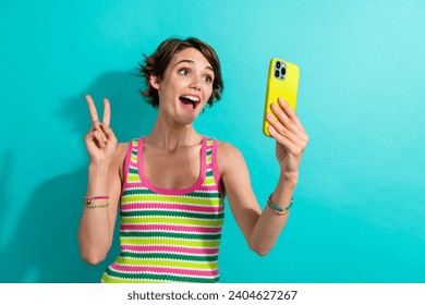 Photo of funky cheerful girl hold smart phone make selfie demonstrate v-sign isolated on teal color background