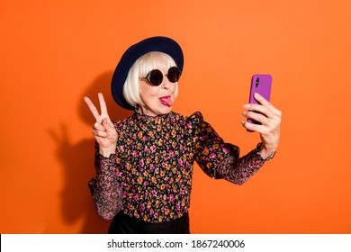 Photo of funky crazy modern hipster grandmother take selfie on phone stick tongue out show v-sign isolated on orange color background