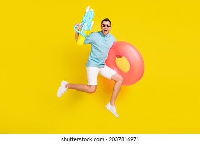 Photo of funky sweet young guy dressed blue t-shirt eyewear jumping holding inflatable rings water gun isolated yellow color background