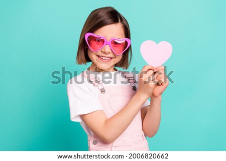 Photo of funny adorable school girl wear pink overall glasses smiling holding little heart isolated teal color background