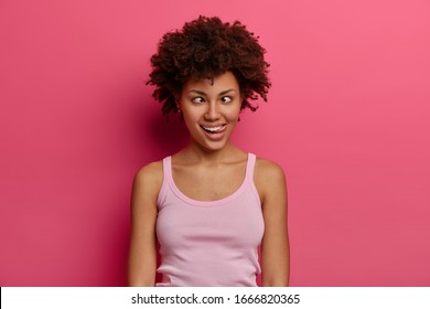 Photo of funny comic woman makes grimace, squints eyes and sticks out tongue, plays fool, has fun alone, being crazy and silly, dressed in casual wear, poses against rosy background. Easy lifestyle