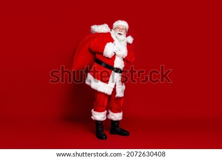 Photo of funny excited man pensioner dressed santa claus costume carrying big christmas sack smiling isolated red color background