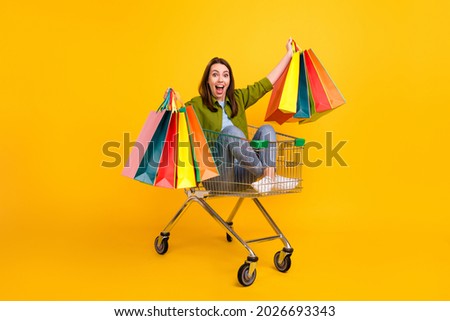 Photo of funny excited young woman dressed green shirt holding bargains riding cart smiling isolated yellow color background