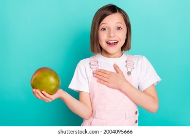 Photo of funny impressed school girl wear pink overall smiling holding huge mango arm chest isolated teal color background